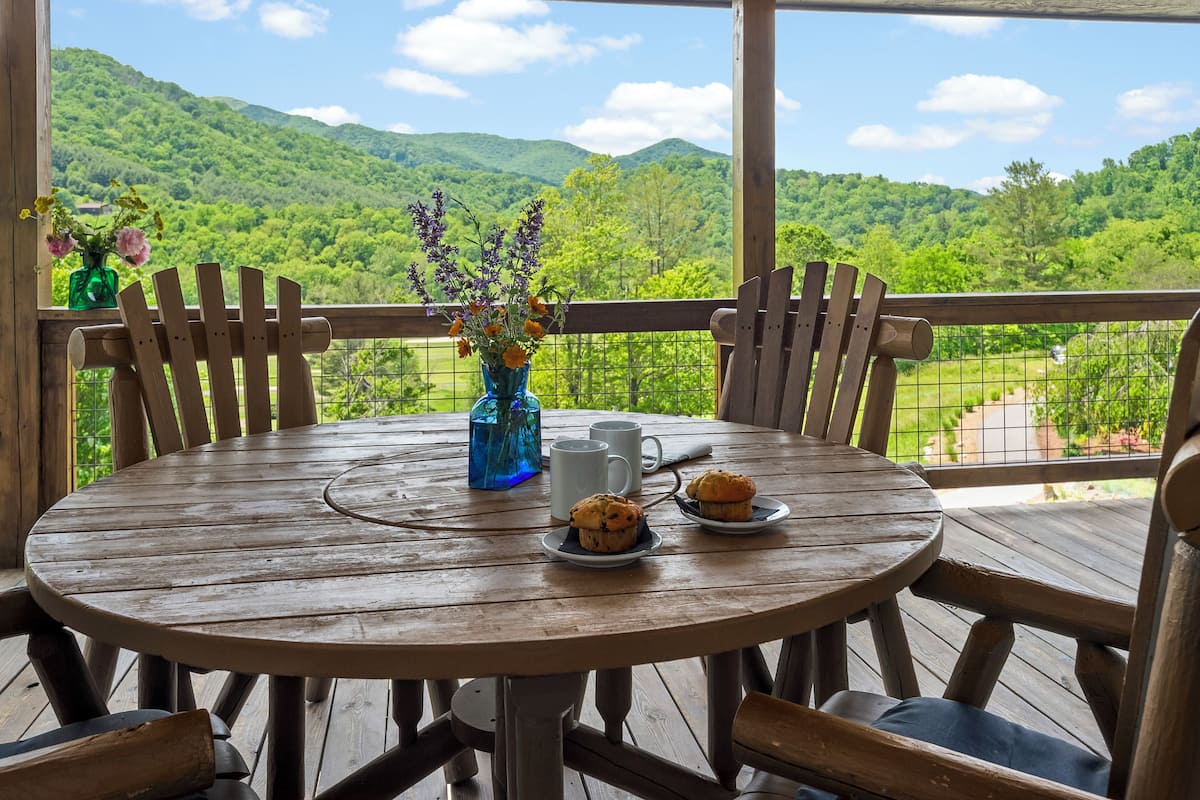 A wooden table with chairs on a balcony, a vase with flowers, and two muffins on plates.