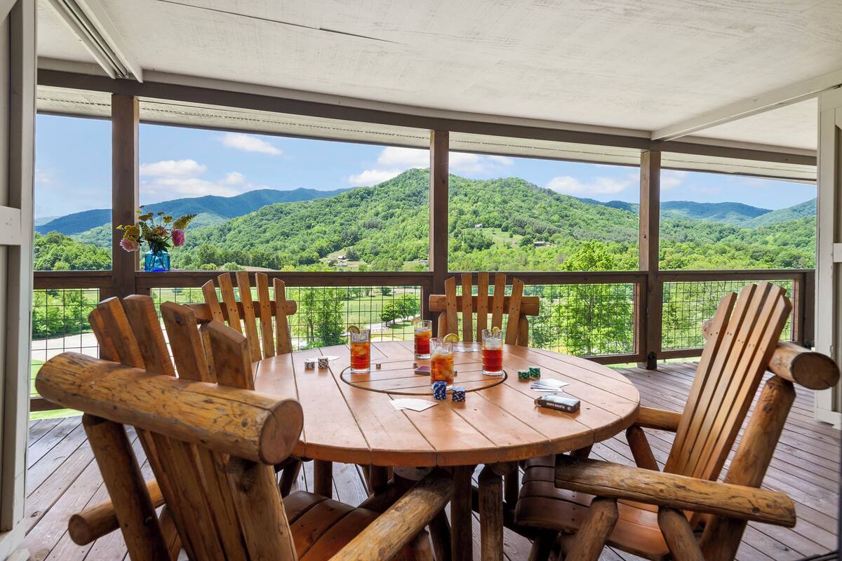 A wooden deck with a round table and chairs, overlooking a lush green landscape with mountains.