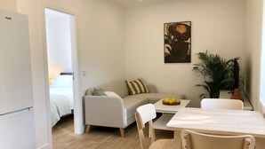 Un espace de vie compact comprenant un coin cuisine, un espace repas avec une table et des chaises, un canapé avec des coussins, un tableau au mur et une plante en pot.