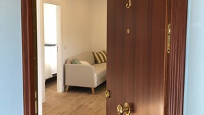 Une porte en bois, dotée d’une poignée en laiton et portant le numéro « 5 », donne sur une pièce où se trouvent un canapé beige, un coussin à motifs et un lit recouvert d’une couverture blanche.