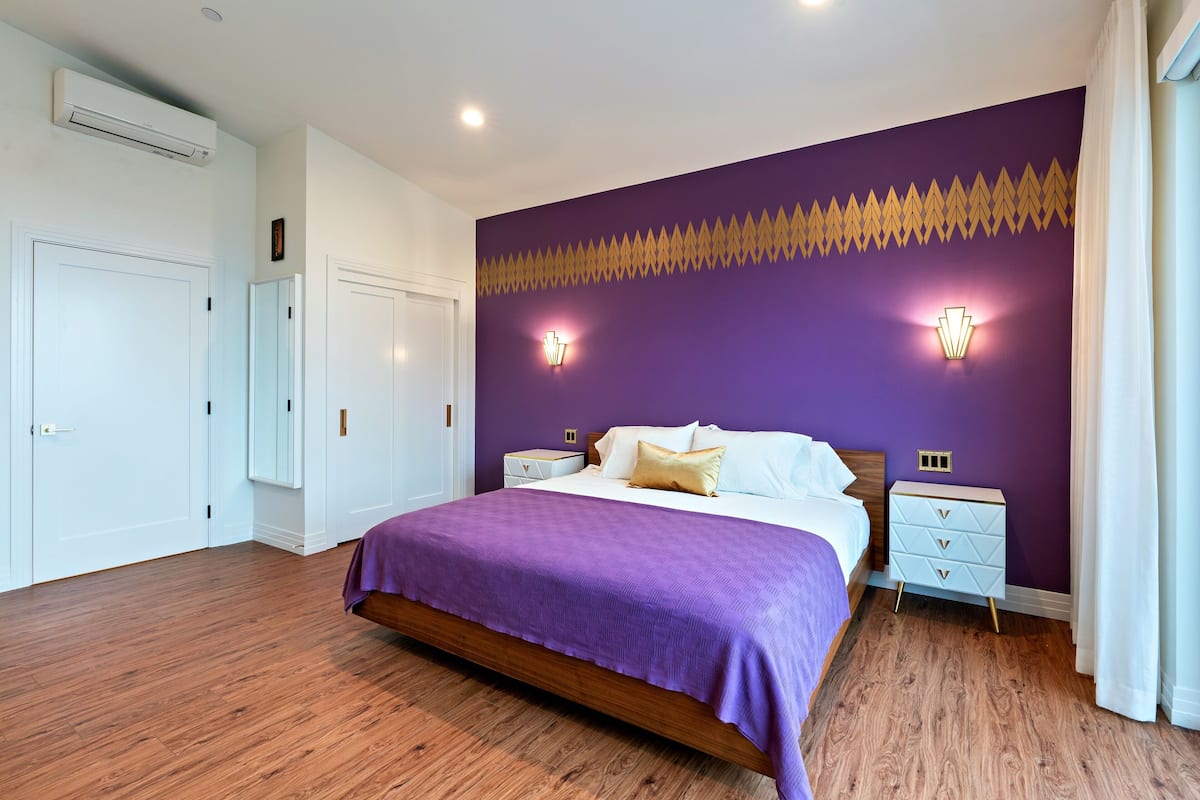 A bedroom with a purple accent wall, a wooden floor, a bed with a purple bedspread, two bedside tables with lamps, and a white dresser.