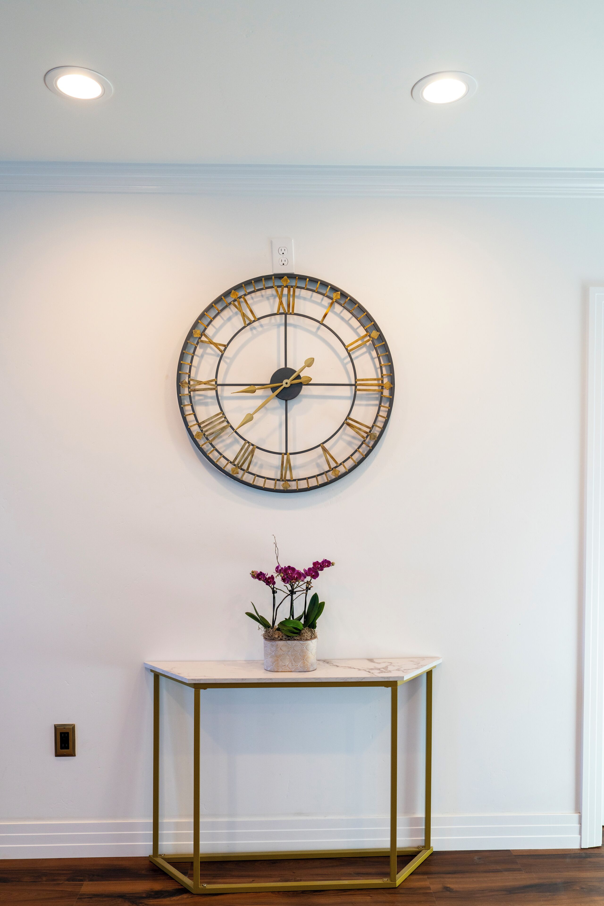 A wall-mounted clock with Roman numerals, a small table with a potted plant, and a white door.