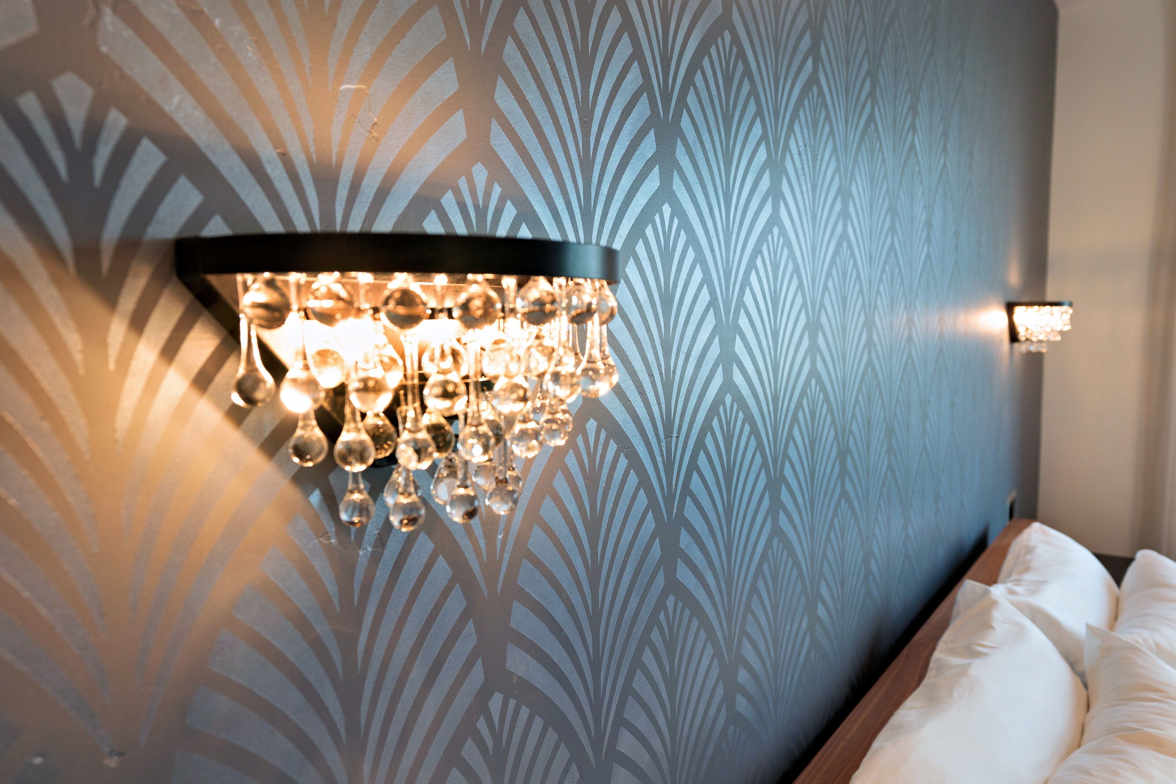 Una lámpara de pared con bombillas esféricas, un papel tapiz decorado y una cama con ropa de cama blanca.