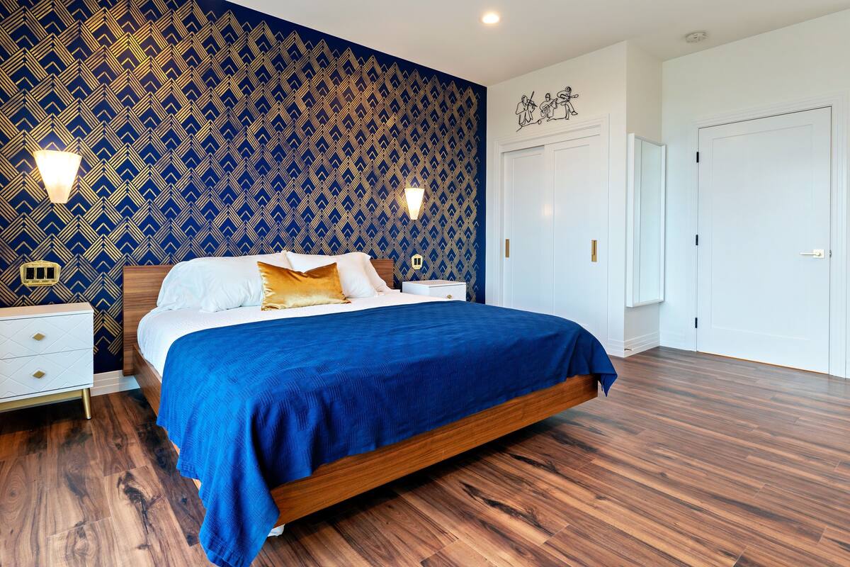 A modern bedroom with a patterned wallpaper, a large bed with a blue bedspread, wooden flooring, and white furniture.