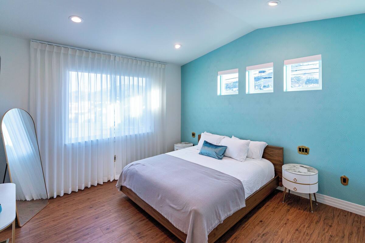 A bedroom with a bed, white curtains, a wooden floor, and a blue accent wall.