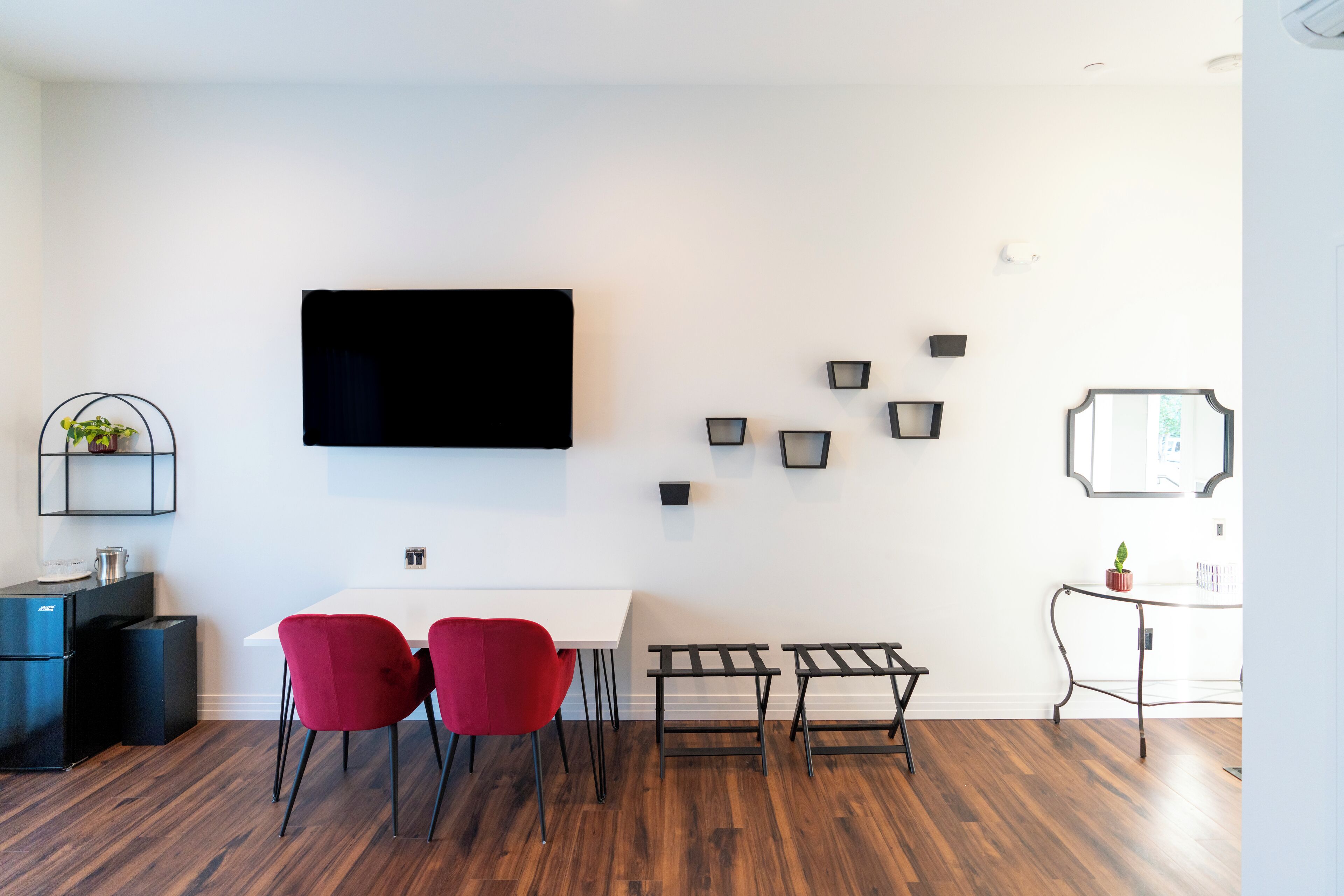 Uma sala de estar moderna com televisão de tela plana na parede, uma mesa de jantar com cadeiras vermelhas, um pequeno sofá preto e uma cafeteira.