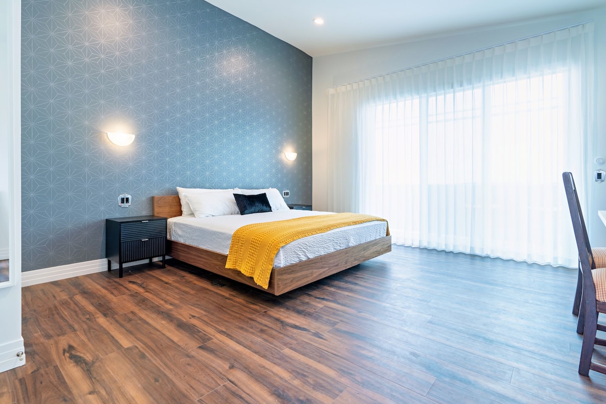 A bedroom with a large bed, wooden flooring, and a window with sheer curtains.