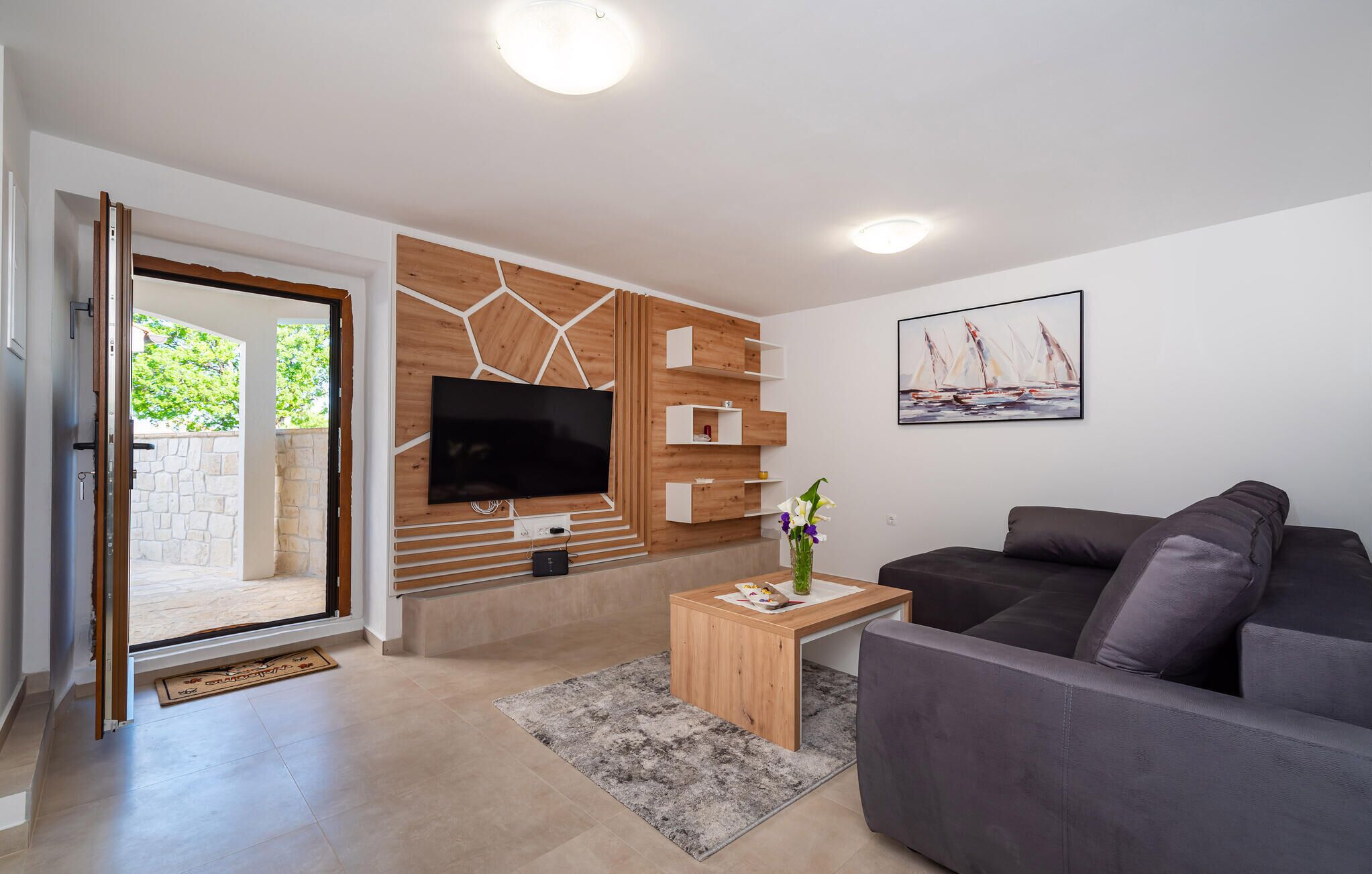 A modern living room with a flat-screen TV mounted on a wooden wall, a gray sofa, a small wooden side table with a vase of flowers, and a painting of sailboats on the wall.