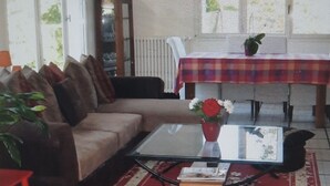 A living room with a sofa, a coffee table with a book, a red rug, a table with a plaid tablecloth, and a plant. - Maison Lumineuse et Spacieuse en Forêt (Buthiers)