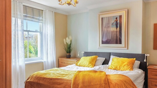 Une chambre à coucher avec un lit, une table de chevet, une lampe, une fenêtre avec des rideaux et un tableau au mur.