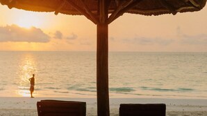 Een strandscène met twee houten ligstoelen onder een rieten parasol, met uitzicht op de oceaan bij zonsondergang.