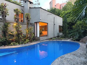 En modern byggnad med en swimmingpool, omgiven av frodig grönska och höga byggnader i bakgrunden.
