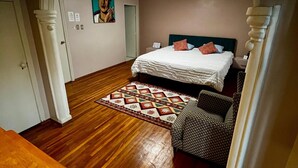 Un dormitorio con suelo de madera, una cama con colcha estampada, una pequeña silla y un cuadro en la pared.
