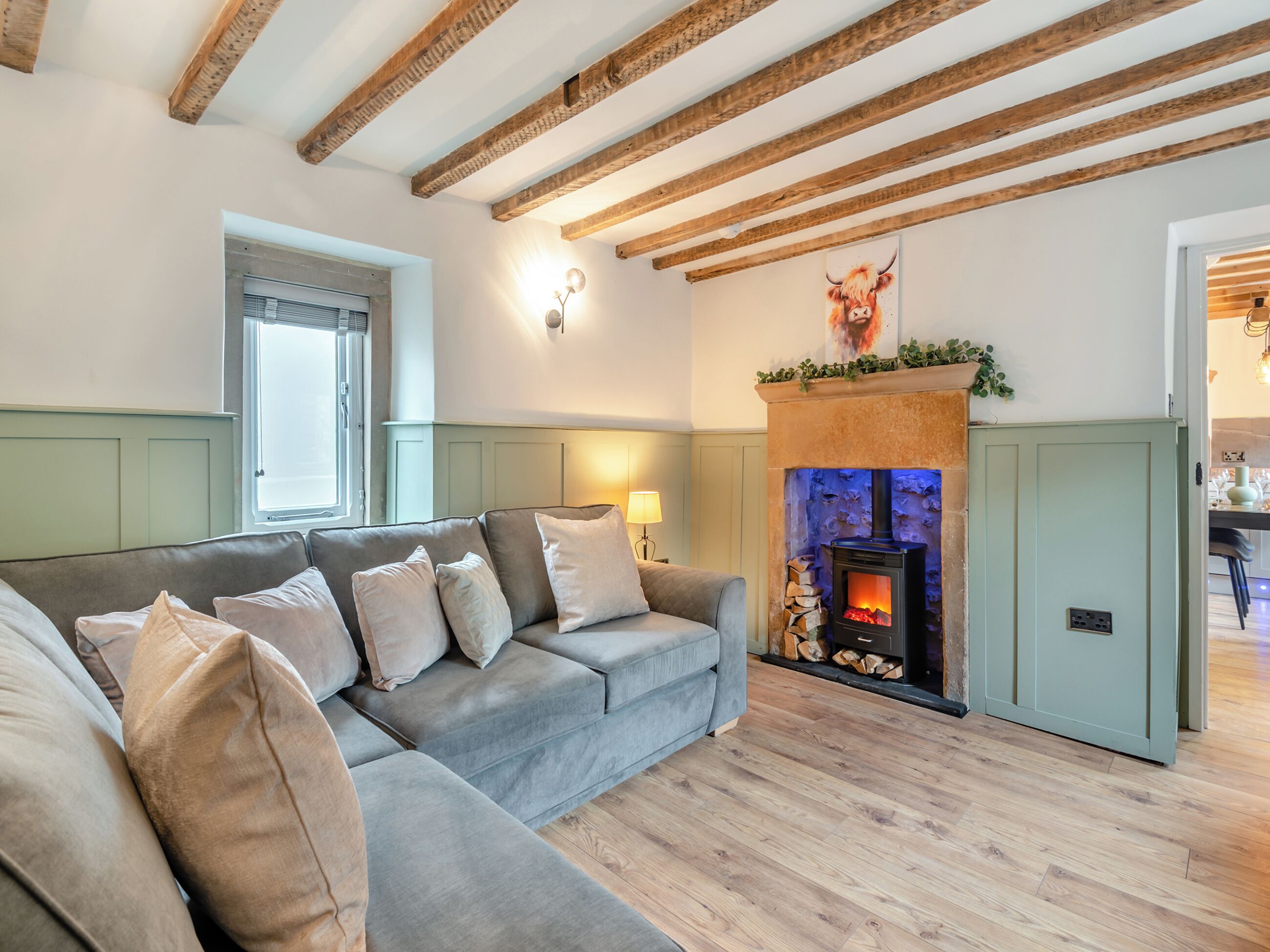 A cozy living room with a grey sofa, a fireplace, and exposed wooden beams.