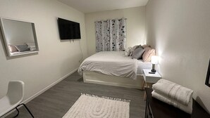 Une chambre à coucher avec un lit, une table de chevet avec une lampe, une chaise, un miroir et une fenêtre avec des rideaux.
