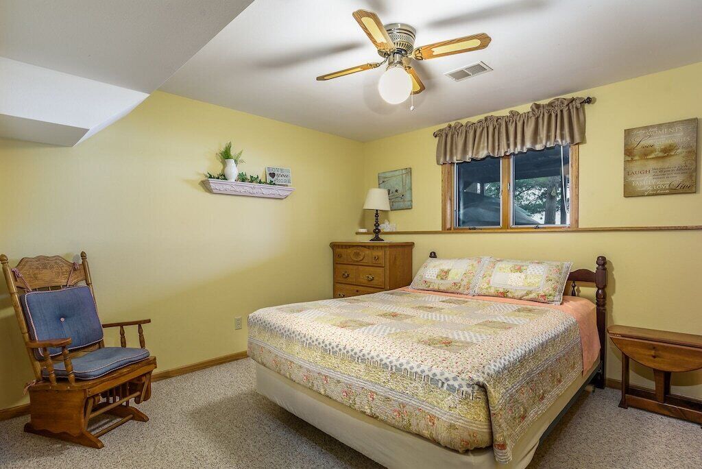 A bedroom with a bed, a wooden chair, a nightstand, a dresser, a ceiling fan, a window with curtains, and a wall-mounted shelf with plants.
