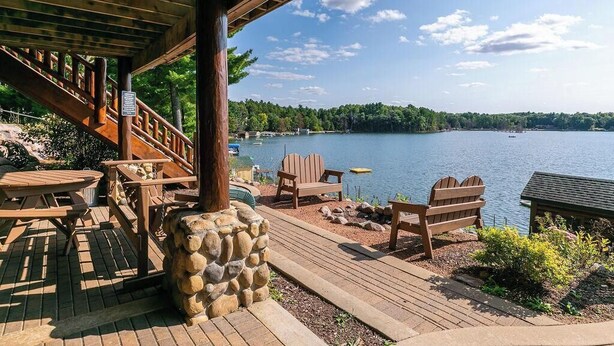 Une terrasse en bois, agrémentée de chaises Adirondack et d’un jardinière en pierre, qui offre une vue sur un lac où l’on peut apercevoir des bateaux, ainsi que sur une rive bordée d’une forêt.