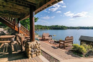 A wooden deck with Adirondack chairs and a stone planter, overlooking a lake with boats and a forested shoreline.