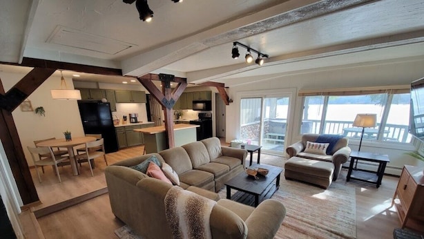 A spacious living area with a large sofa, a coffee table, and a dining table. There is a kitchen in the background with a refrigerator and cabinets. - Amazing view, hot tub, sauna, fireplace, pet friendly, pier, kayaks, paddleboard (Wautoma)