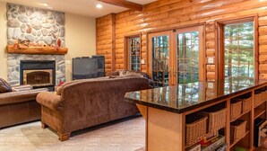 A cozy log cabin interior with a stone fireplace, a leather sofa, a television, and a large window with a view of trees. - Free nite w/wknd offseason;logcabin lake hottub kayak paddleboard pier fireplace (Wild Rose)