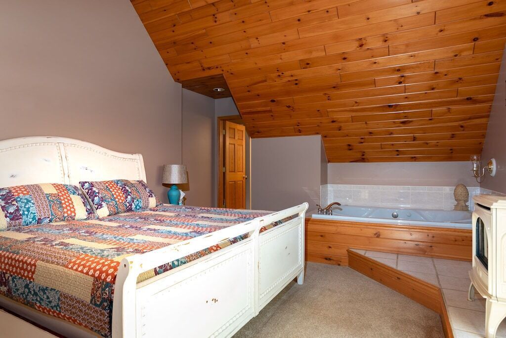 A bedroom with a wooden ceiling, a white bed with a colorful quilt, a wooden dresser, a wooden bench, and a bathtub.