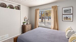 Ein Schlafzimmer mit Bett, Nachttisch, Kommode, einem Fenster mit Vorhängen und Wanddekoration.