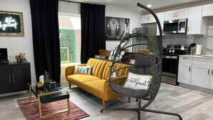 A modern living room with a yellow sofa, a glass coffee table, and a hanging chair. There is a kitchen area with white cabinets and a microwave.