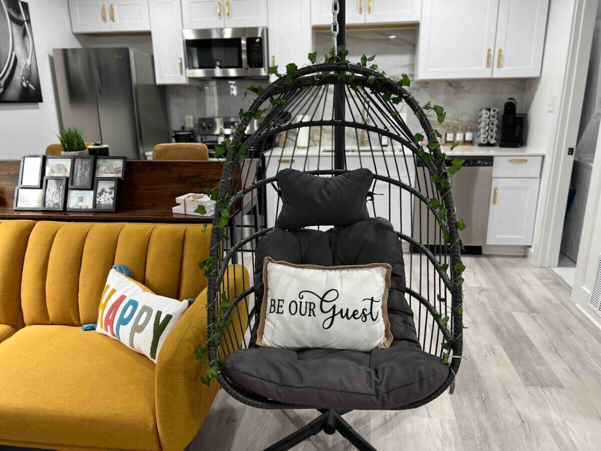 A modern kitchen with white cabinets, stainless steel appliances, and a hanging chair with a 'BE OUR GUEST' pillow.