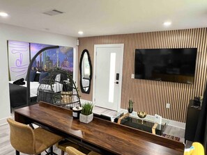 A modern living room with a wooden dining table, a sofa, a TV mounted on the wall, and a cityscape painting.