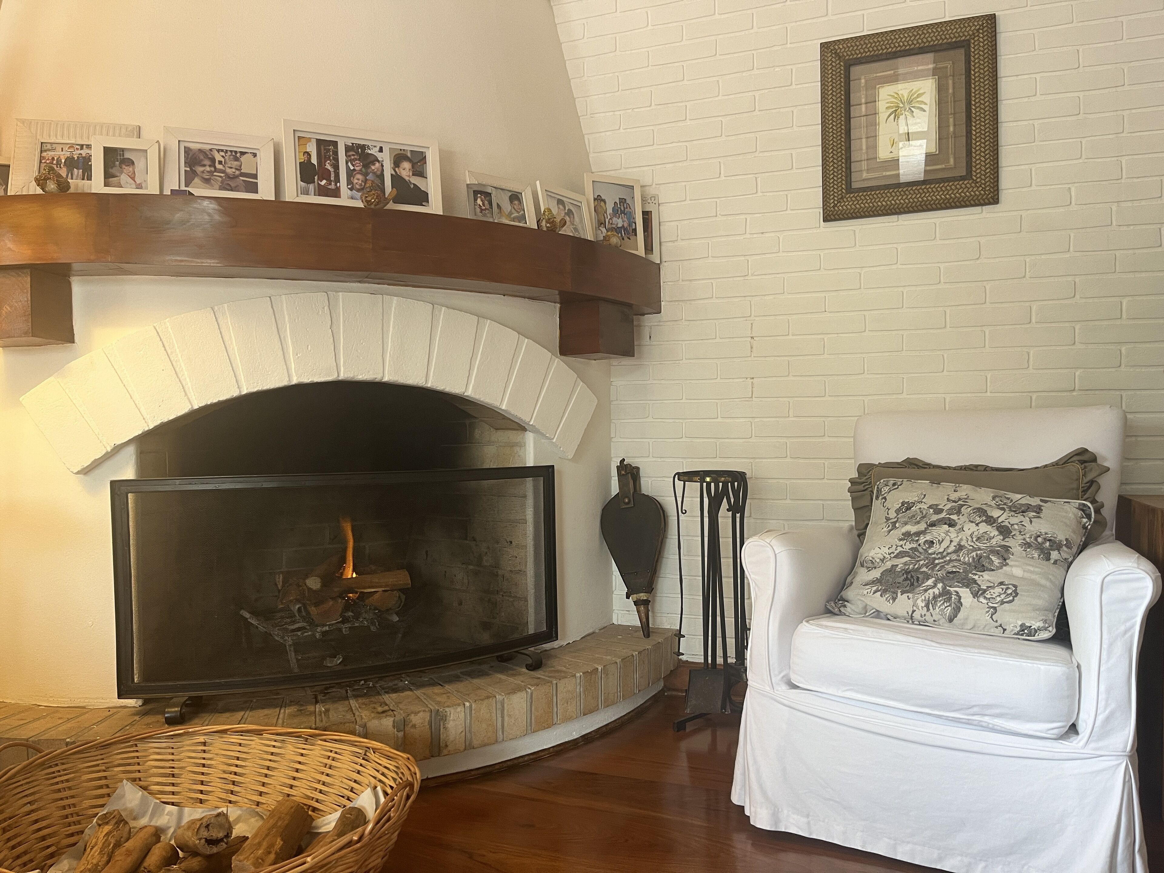 A living room with a fireplace, a white armchair with a floral pillow, and a basket of wood logs.