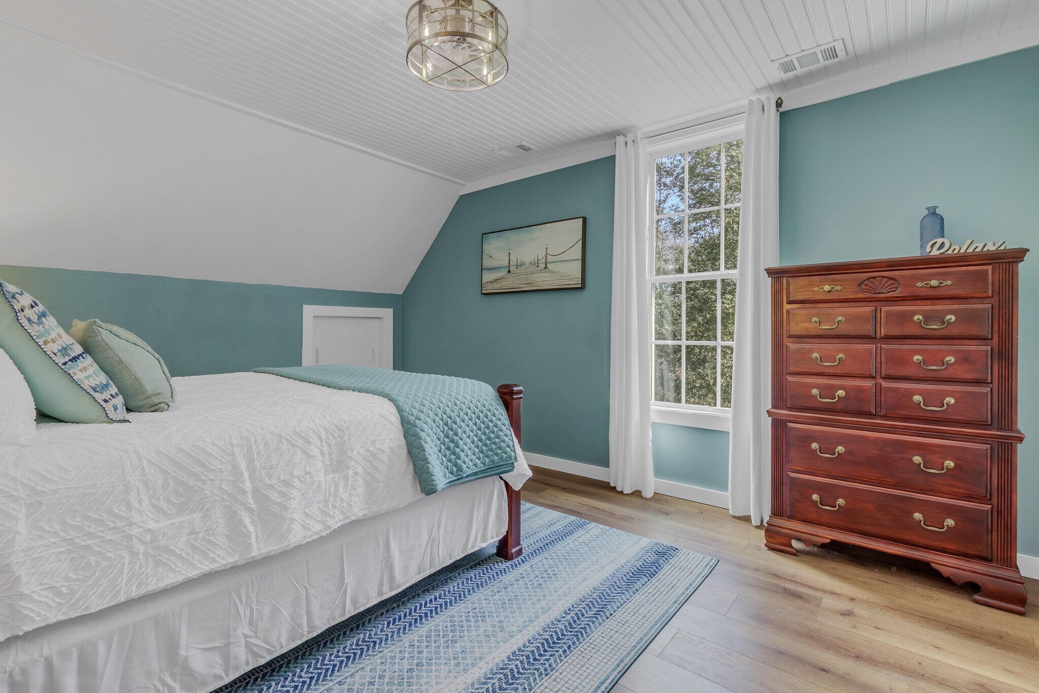 A bedroom with a bed, a dresser, a window, and a painting on the wall.