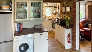 Eine kompakte Küche mit Waschmaschine, ein Essbereich mit rot-ledriger Couch und Blick auf einen Garten durch ein Fenster.