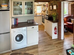 Uma cozinha compacta com máquina de lavar, uma área de refeições com sofá de couro vermelho e vista para um jardim pela janela.