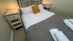 Un lit bien fait, recouvert d’une couvre-lit à motifs et agrémenté d’un seul oreiller, est encadré par deux tables de chevet sur lesquelles reposent des lampes, dans une pièce simple aux murs unis.