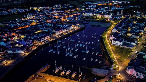 Vue aérienne d’une ville côtière de nuit, avec un port de plaisance rempli de bateaux, entouré de bâtiments résidentiels et d’une église imposante.