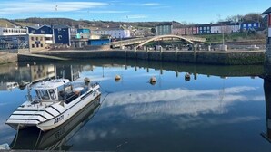 Une rivière paisible, avec un bateau amarré sur la rive gauche, un pont piétonnier au centre et des bâtiments le long de la berge.