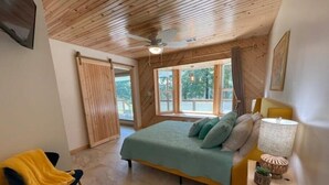 Quarto com cama, cadeira, luminária e janela com vista para vegetação.