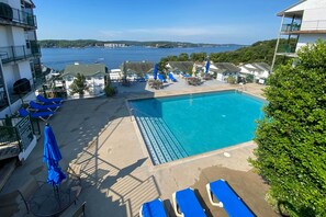 Una piscina con sillas reclinables azules y sombrillas, rodeada de edificios con balcones que dan a un cuerpo de agua.