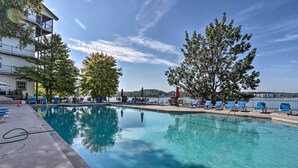 En swimmingpool med kristallklart blått vatten, omgiven av solstolar och träd, med en byggnad i bakgrunden.