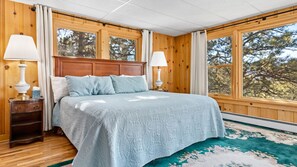A bedroom with a wooden headboard, two bedside tables with lamps, a large bed with a light blue bedspread, and a view of trees through the windows. - Family-Friendly 4BR Home w/ Views – Minutes to Town (Estes Park)