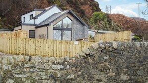 A two-story house with a stone wall in the foreground, a wooden fence, and a hillside in the background. - 2 bedroom accommodation in Lochranza (Lochranza)