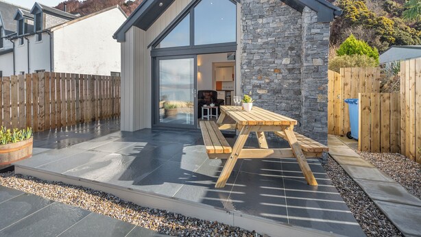 A modern house with a stone exterior, a large glass door, and a patio with a wooden table and chairs. - 2 bedroom accommodation in Lochranza (Lochranza)