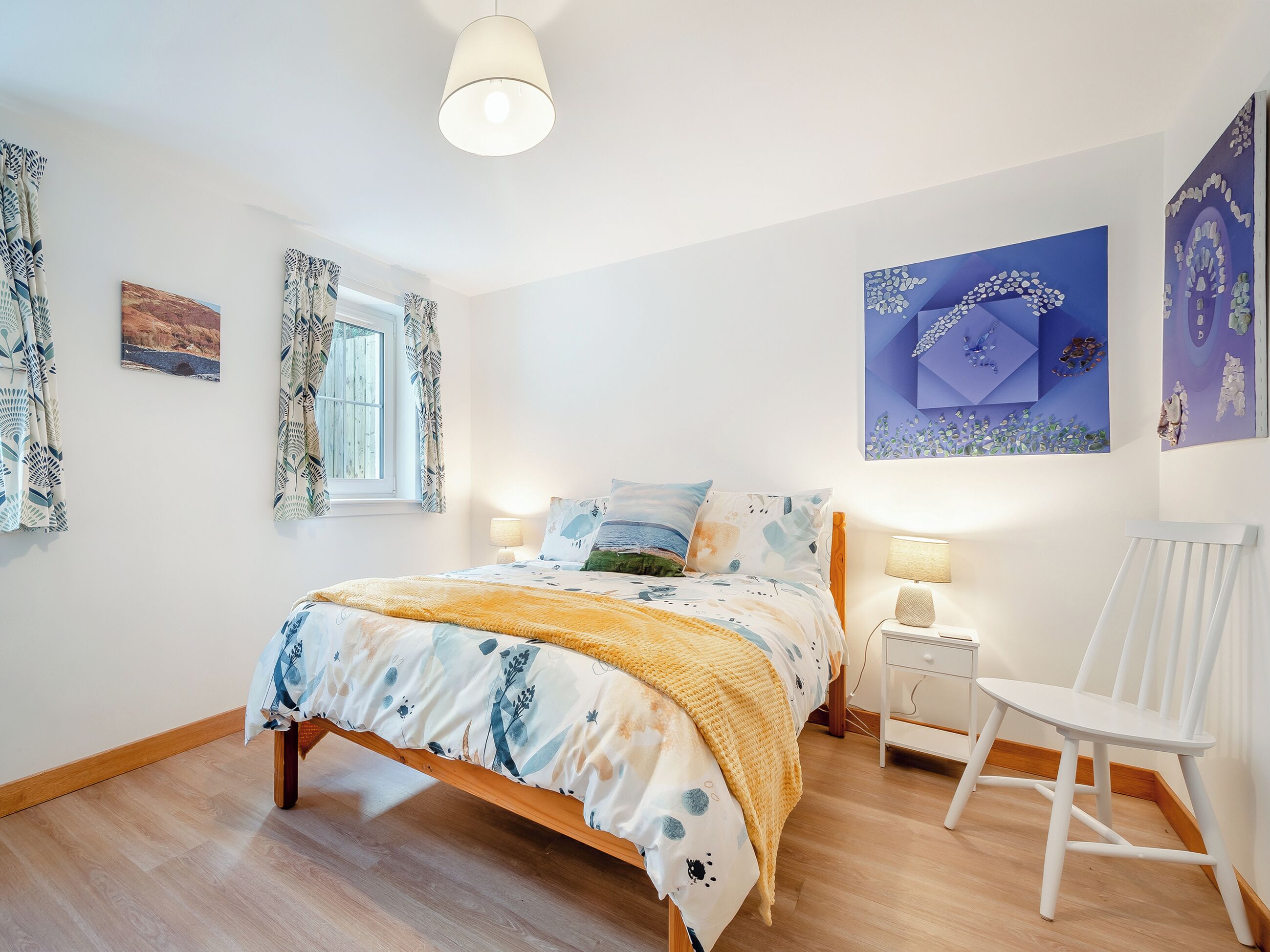 A bedroom with a bed, a window with curtains, a bedside table with a lamp, and a chair.