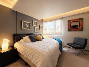 A modern bedroom with a bed, bedside table, chair, and artwork on the walls.