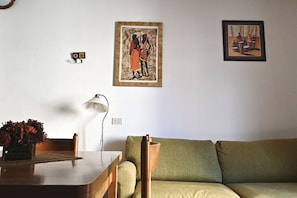 A living room with a wooden table, a green sofa, a lamp, and two framed artworks on the wall.