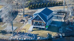 An aerial view of a modern house with a large deck, a covered outdoor area, and a stone wall by the water.