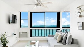 Un salón moderno con un ventanal grande con vistas al océano, un ventilador de techo, un televisor de pantalla plana y una decoración minimalista.