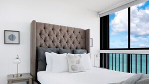Un dormitorio con cabecera capitonada, ropa de cama blanca, vista al océano a través de un balcón y una mesita de noche con lámpara.