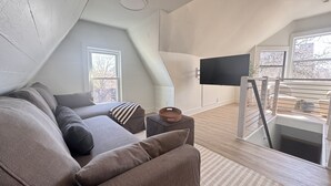 A modern loft bedroom with a sloped ceiling, a large window, a flat-screen TV, a staircase, and a cozy seating area with a sofa and cushions.