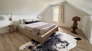 A bedroom with a sloped ceiling, a bed with striped bedding, a cowhide rug, a small wooden stool, a wall-mounted lamp, and a window with curtains.